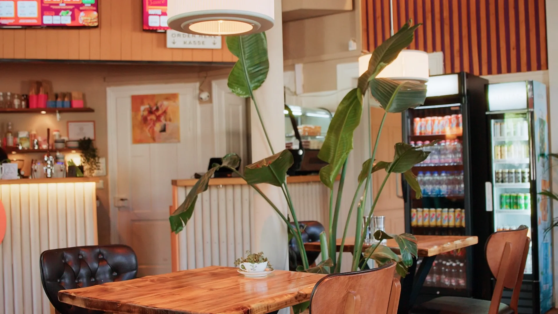 Inside DÄ BURGER: wooden tables, a big plant, the counter with the menu boards above it and fridges full of drinks