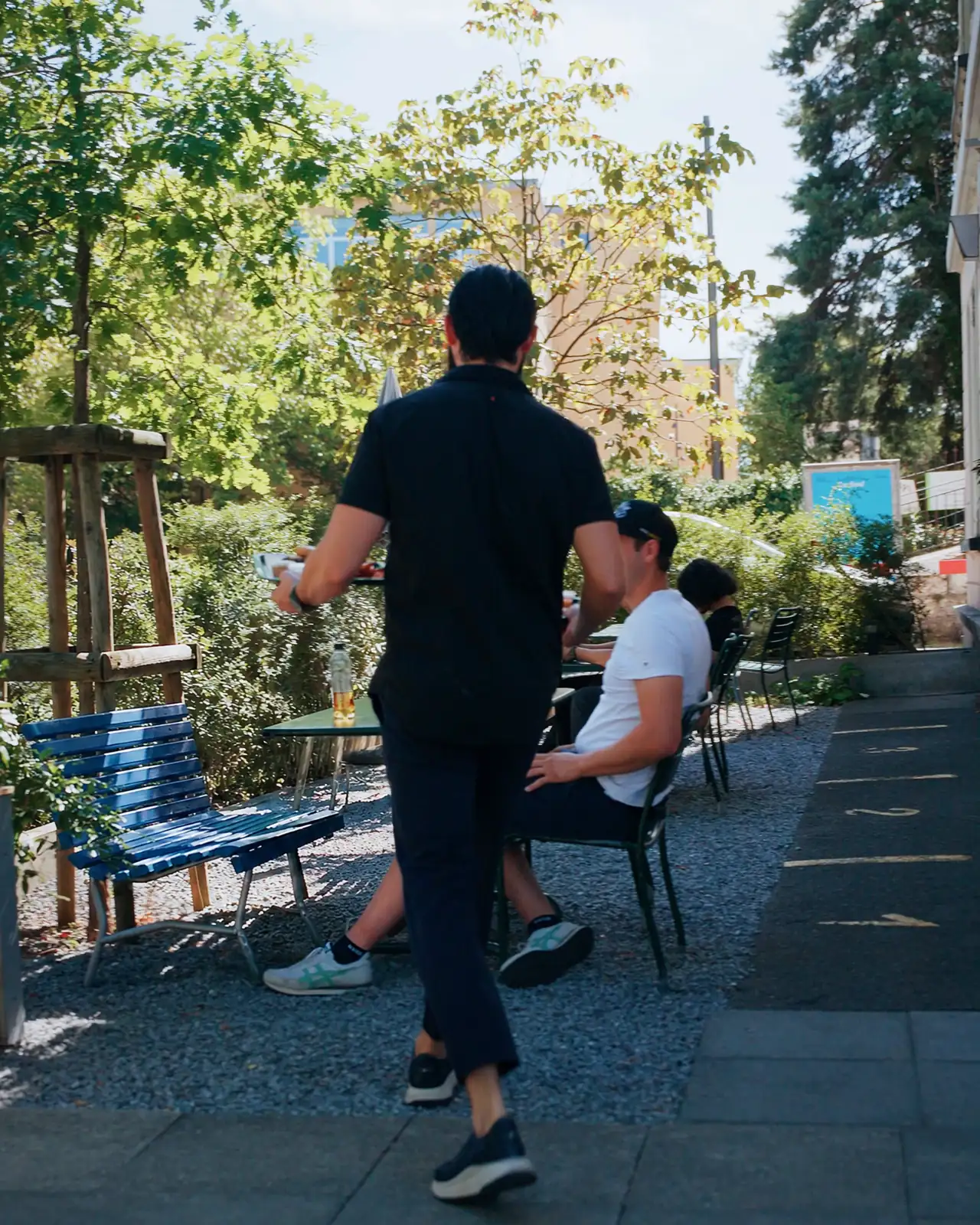 Our garden terrace in summer: tables on gravel under the trees, a blue bench on the left, a guest at a table, a staff member bringing his order